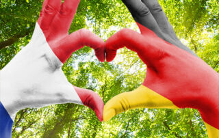 Zwei Hände formen ein Herz vor grünem Blätterdach, bemalt in den Nationalfarben von Frankreich und Deutschland – ein Symbol für deutsch-französische Freundschaft und Verbundenheit.